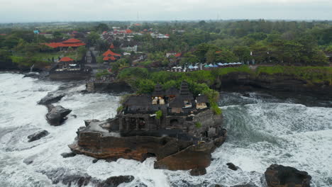 Tilting-aerial-shot-flying-away-from-empty-Tanah-Lot-temple-during-dangerous-weather-in-Bali,-Indonesia.-Dangerous-sea-waves-crashing-into-dark-rocks-with-famous-Hindu-temple