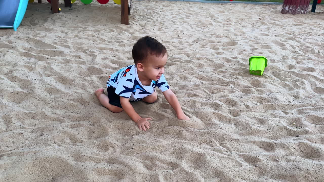 Happy joyful toddler crawls by the white sand on the playground. Adorable child having fun on the walk in summer.