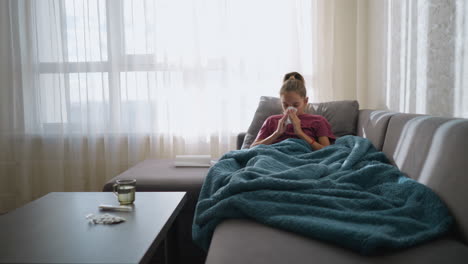 girl in pink suffering from flu lying on couch, sneezing into tissue with medication and glass cup on table, wrapped in blanket, looking exhausted and sick