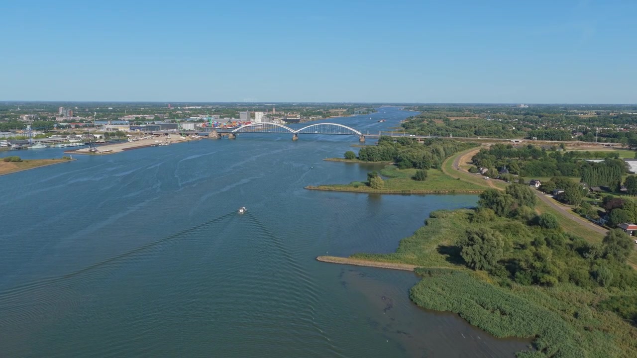 Luftperspektiv af en båd, der sejler på Merwede-floden mod den ikoniske Merwede-bro i Holland under en klar blå himmel.