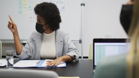 african american businesswoman wearing face mask sitting at desk talking to colleagues in office