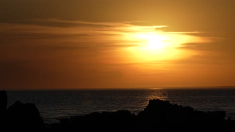 Golden-hour-at-sea,-sun-setting-behind-rocks-with-warm-sky-reflections-on-water,-tranquil-evening