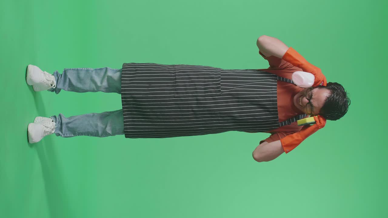 Full Body Of Asian Male Housekeeper With An Apron With A Spray And Kitchen Sponge Then Having A Headache In The Green Screen Studio