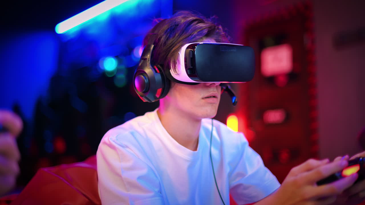 Due amici adolescenti giocano a una console con visore VR e cuffie usando i gamepad, parlano e sorridono mentre sono seduti su pouf. Illuminazione blu e rossa