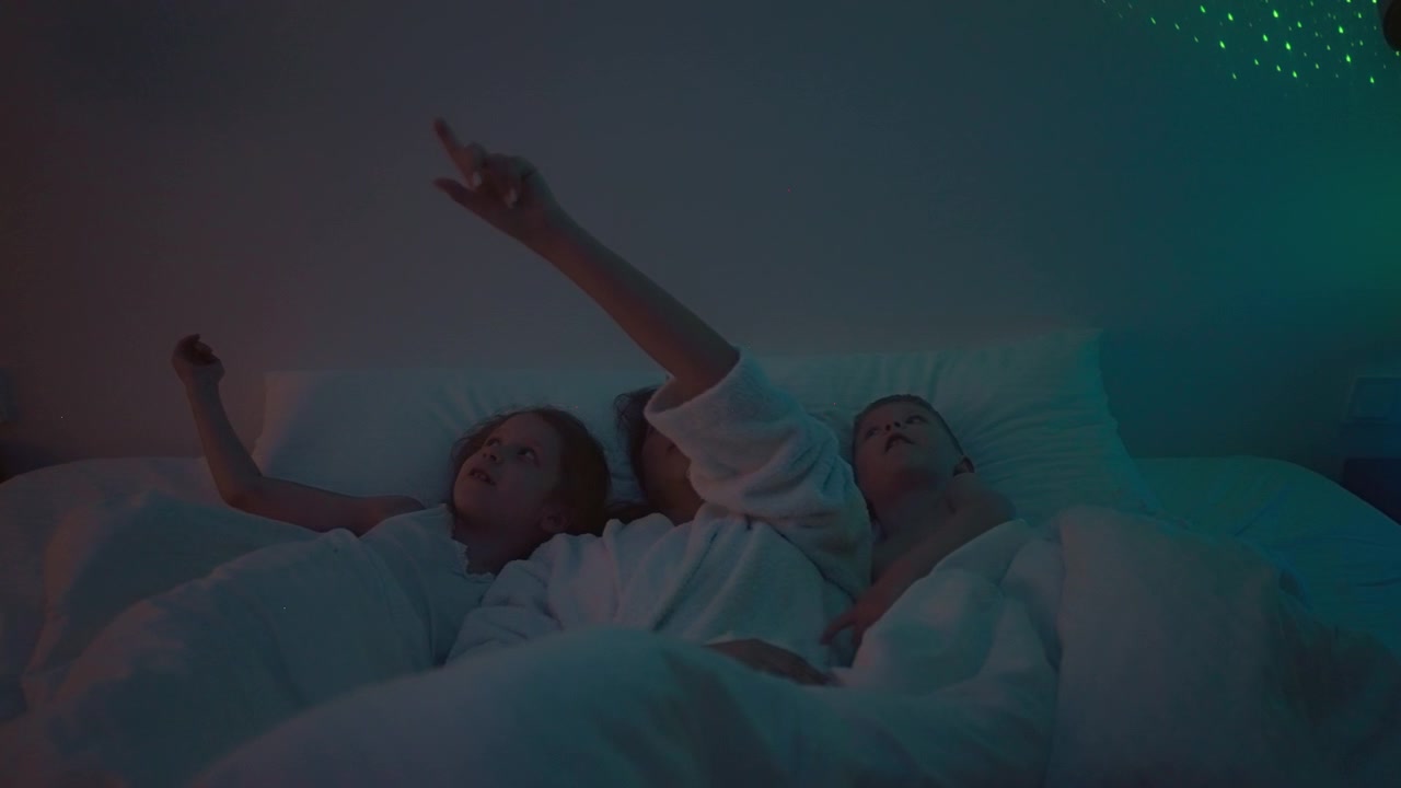 Young woman with kids lie on comfortable bed with projector reflecting colourful stars on wall. Space for children with starry night sky on background