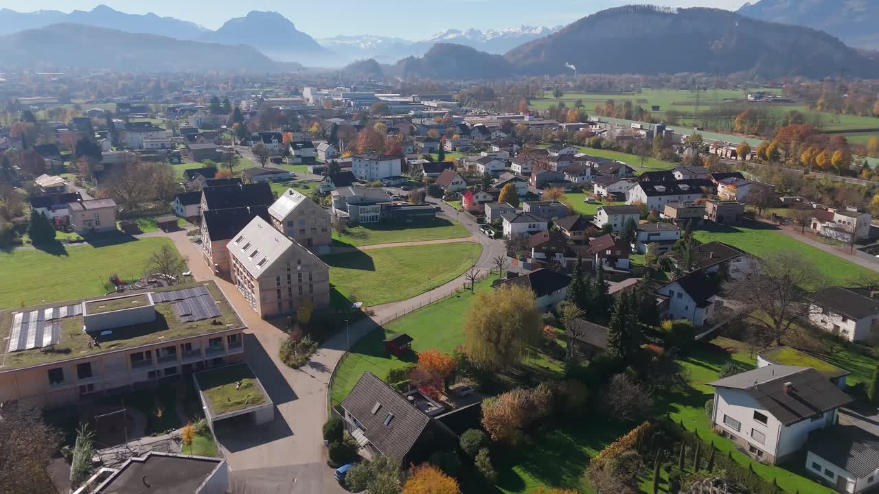 Austrian neighborhood with houses and homes during sunny day in fall season. Silhouette of mountains in distance. Aerial wide shot. Modern buildings with solar panels. Altach, Austria
