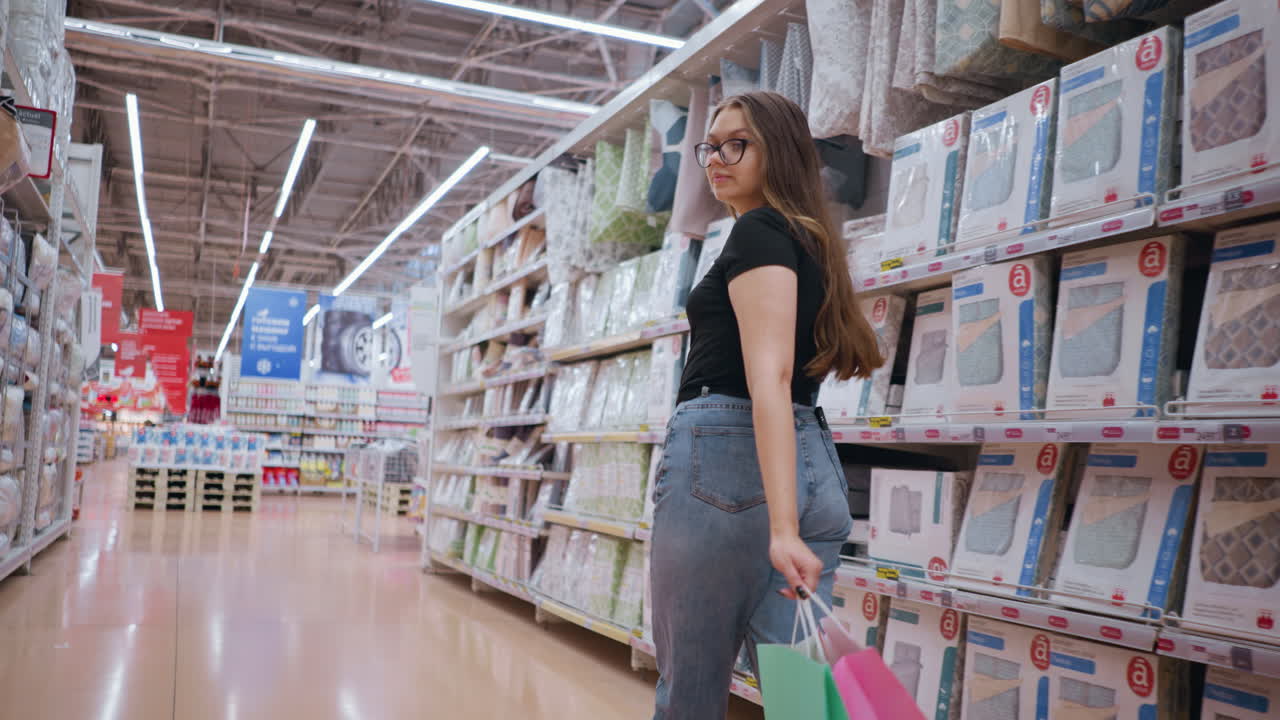 vista trasera de una mujer con camiseta negra y vaqueros caminando por el pasillo de la tienda sosteniendo bolsas de compras, navegando por productos de cama mientras mira a su alrededor, otros compradores se ven en el fondo
