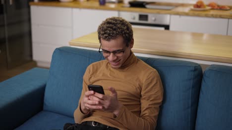 Lockiger-Junger-Mann,-Der-Zu-Hause-Auf-Dem-Sofa-Mit-Seinem-Mobiltelefon-Im-Internet-Surft