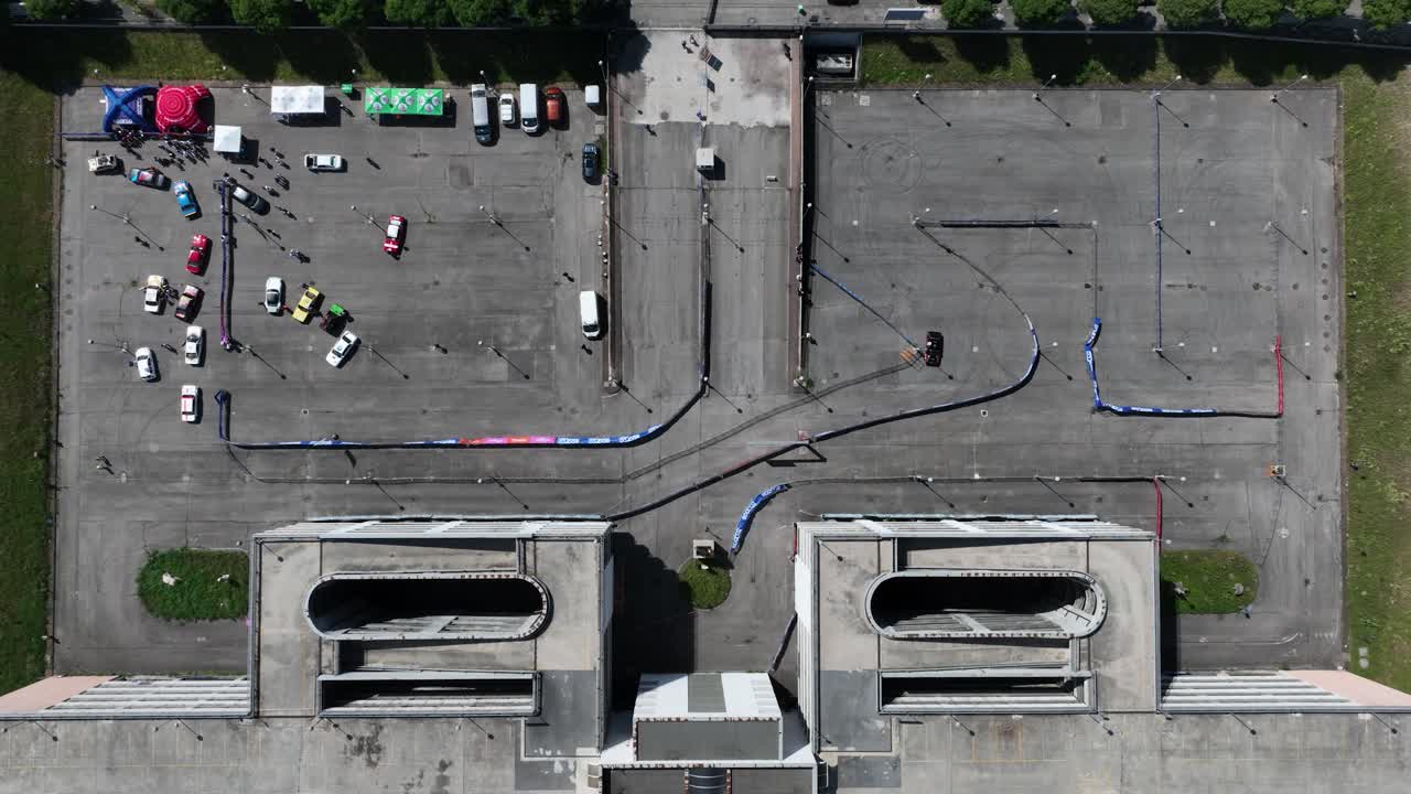 Static top-down drone shot of two black rally cars circling a closed circuit set in a parking lot, passing partly under a multi-level parking garage, with colorful paddock and parked cars