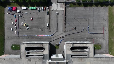 static top-down drone shot of two black rally cars circling a closed circuit set in a parking lot, passing partly under a multi-level parking garage, with colorful paddock and parked cars
