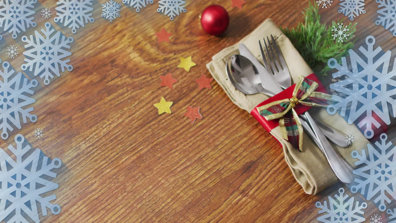 Animation of christmas decorations and cutlery with copy space over snowflakes on wooden background