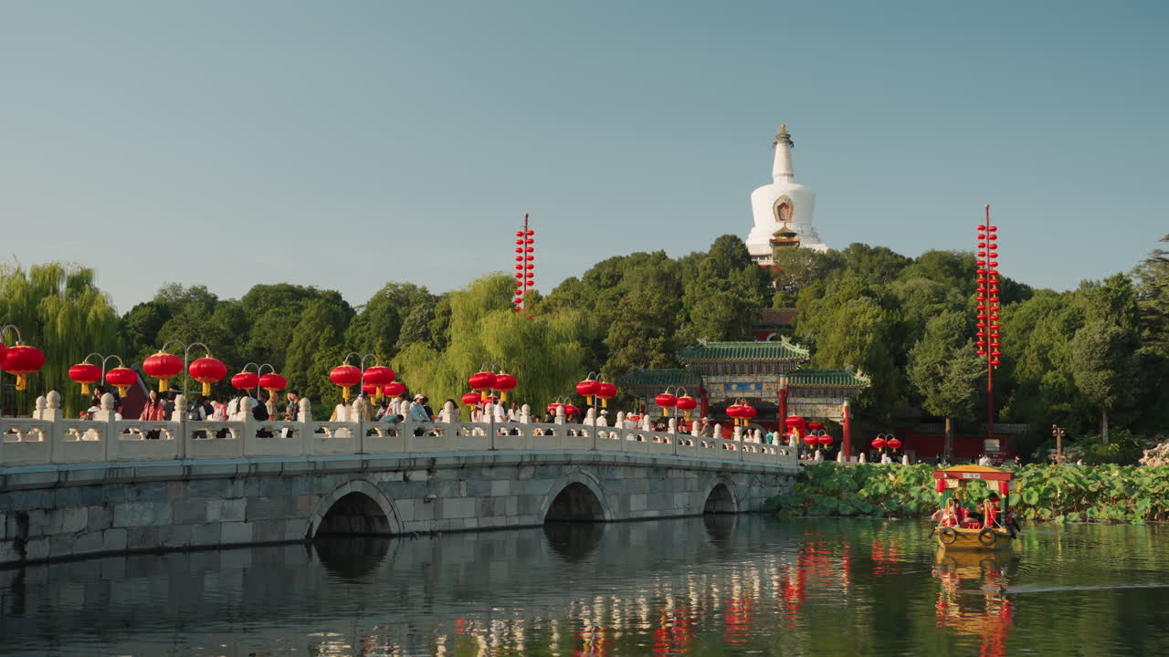 Đám đông trên cầu đá gần ngôi chùa, đèn lồng đỏ và nắng hè rực rỡ, du khách và người biểu diễn địa phương đông nghịt lối đi bên cạnh hồ nước trong như gương, cây cối xanh tốt và tháp stupa trang trí công phu trên đồi, những chiếc thuyền nghi lễ và cảnh quan sống động