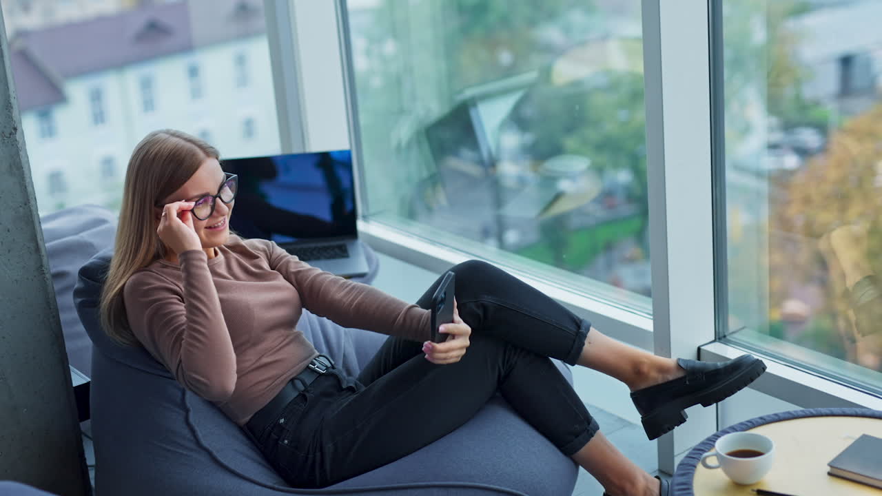 Young lady sitting in bean bag chair fooling around. Woman is taking funny selfie on her phone. High angle view.