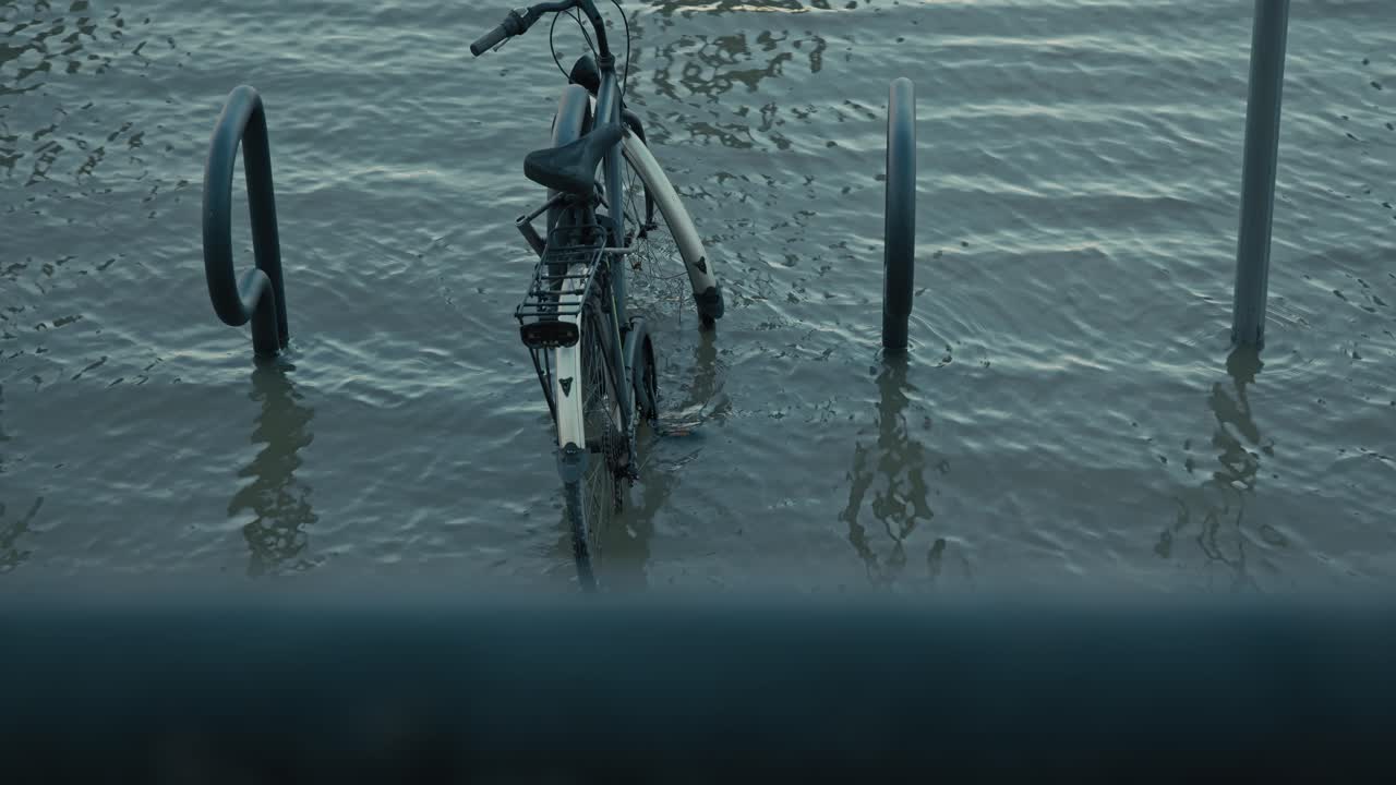 Submerged bike in flooded waters near a bike rack along the riverbank