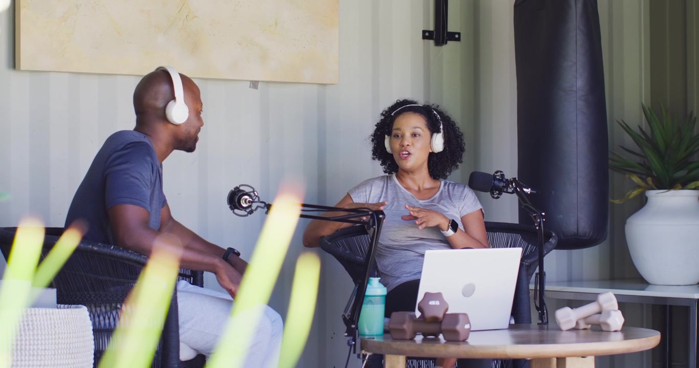 Conduttori afroamericani che registrano un podcast a un tavolo con microfoni, una donna che inizia a parlare, un uomo che risponde.