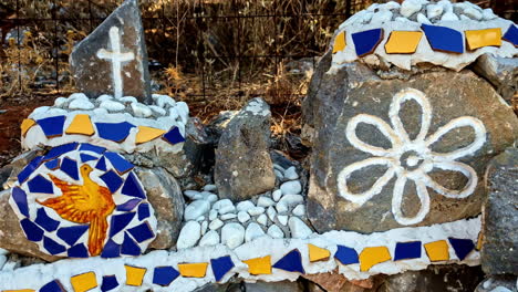 a vibrant mosaic art piece outdoors featuring rocks, a cross, a dove, and floral motifs