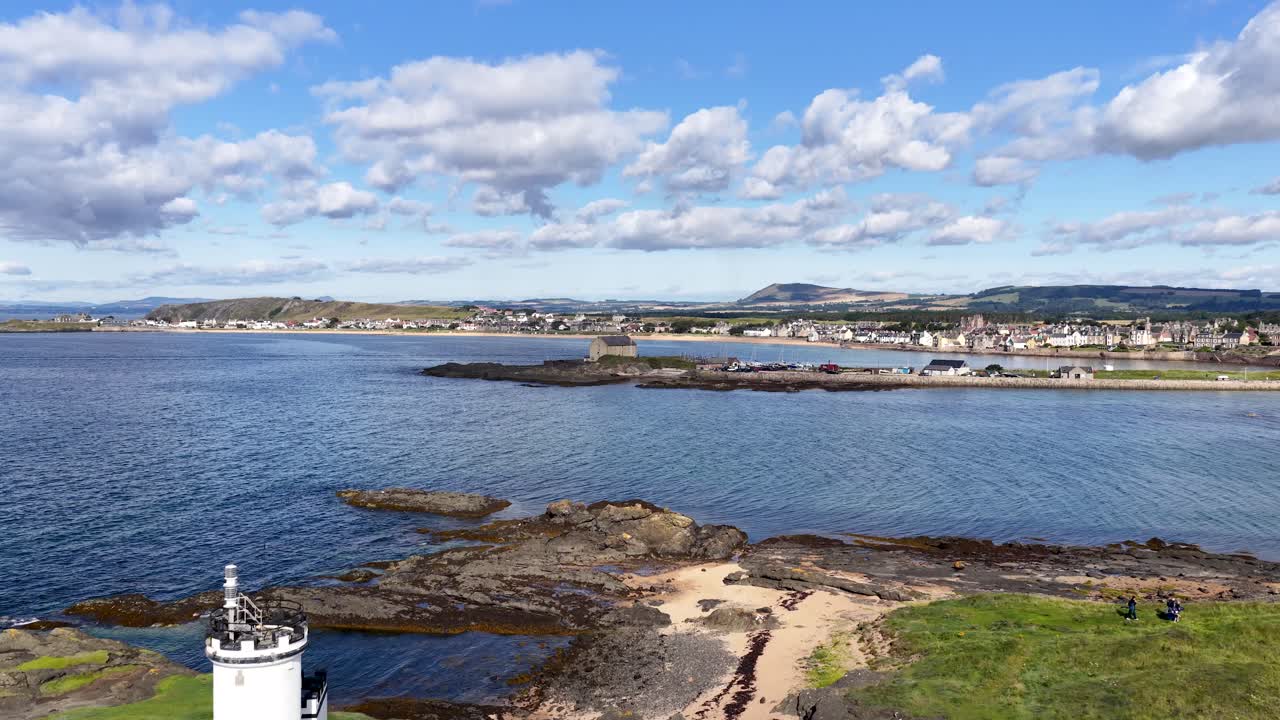 Drone pans above rocky coastline, lighthouse, tents, and seaside village under bright daylight in Fife
