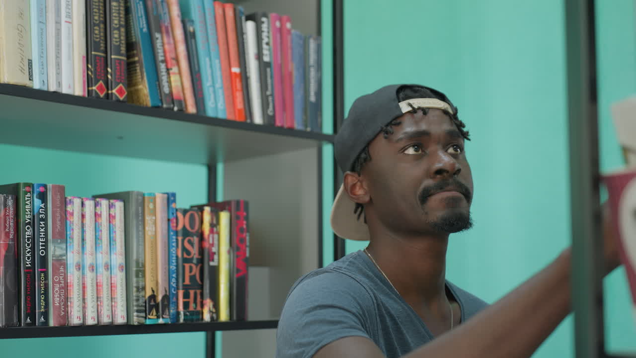 Student wearing stylish casual outfit and backward cap standing between tall library shelves holding book in one hand and smiling warmly in bright modern interior filled with colorful books