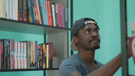 student wearing stylish casual outfit and backward cap standing between tall library shelves holding book in one hand and smiling warmly in bright modern interior filled with colorful books