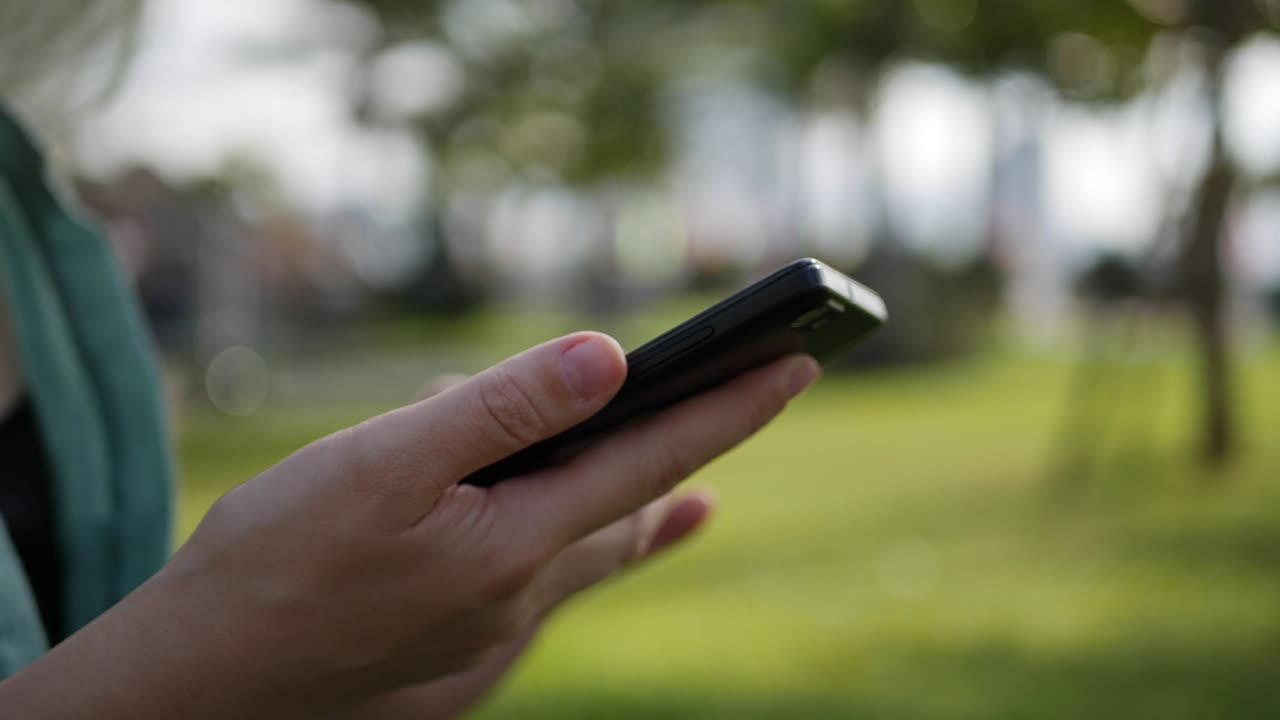 초록색 셔츠를 입고 긴 금발 머리카락을 가진 젊고 아름다운 은 여자가 바닥에 앉아 있으며 공원의 배경에서 스마트폰을 사용합니다.