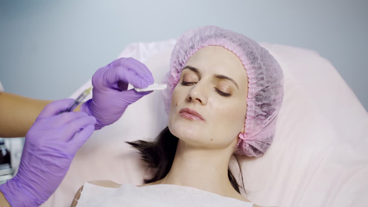 A beautiful woman is lying on a couch at a reception by a beautician. The doctor makes beauty injections to the lips.