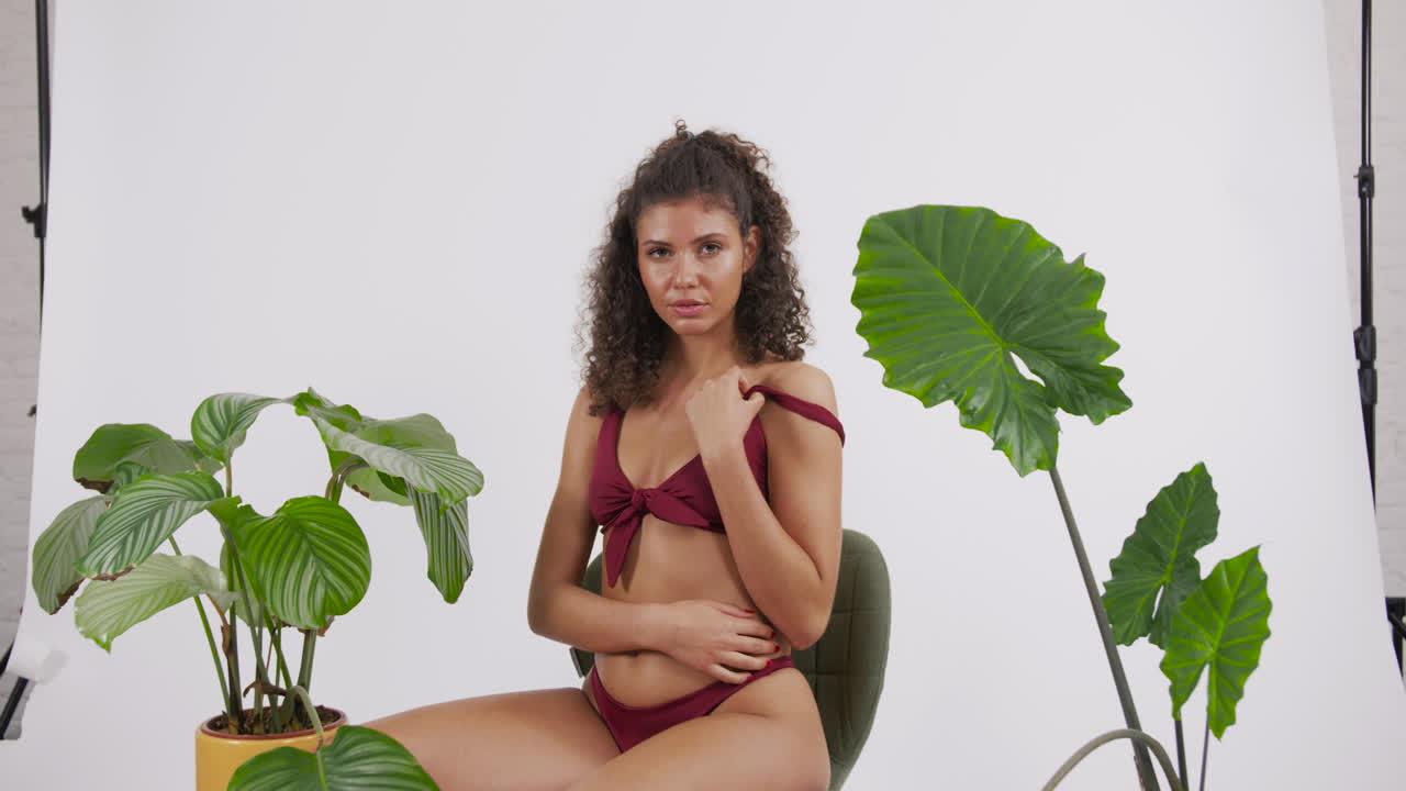 Dark curly haired and tanned model posing on a chair with a red bikini suit, surrounded by plants, shot in a photo studio