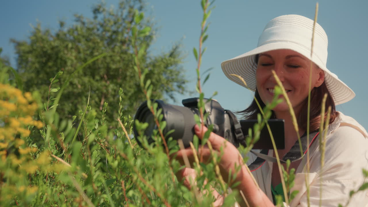 Smiling female tourist in white sunhat squats among tall wild plants holding camera joyfully while filming vibrant flowers in green meadow on clear summer day under bright blue sky