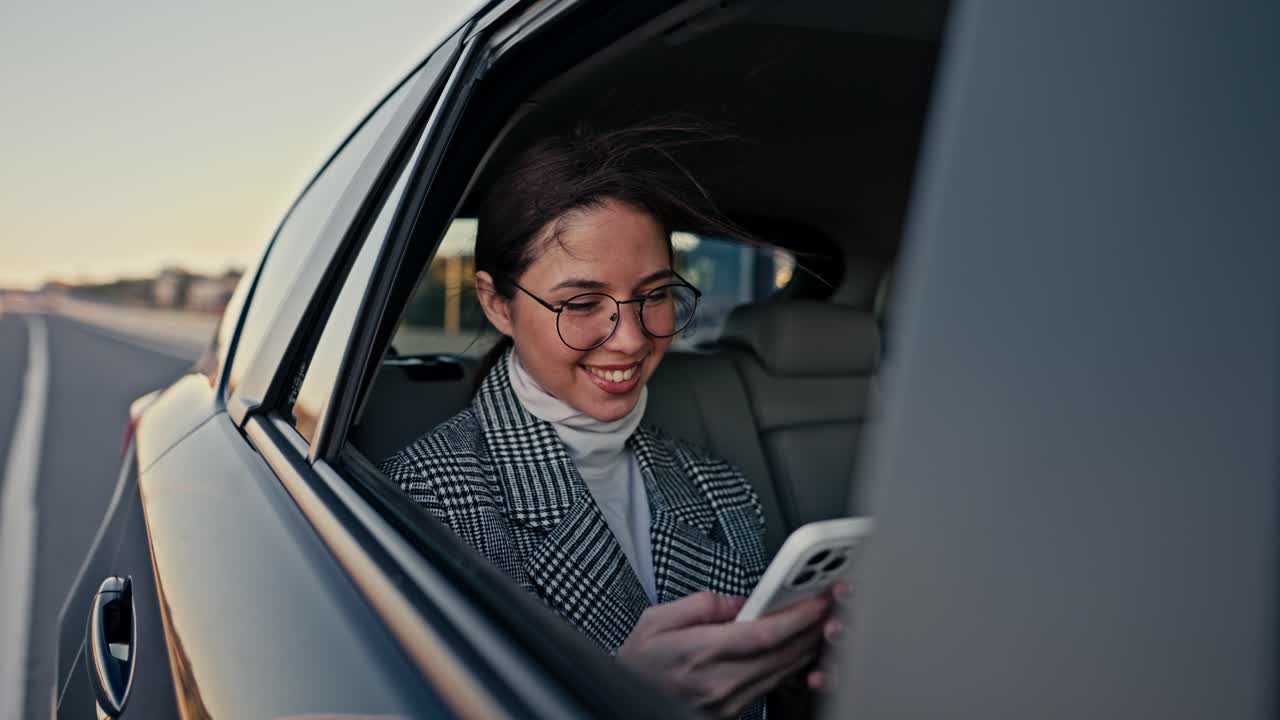 vista exterior de una feliz chica morena con gafas redondas mujer de negocios se comunica en un teléfono blanco y mira por la ventana durante su viaje en un coche moderno