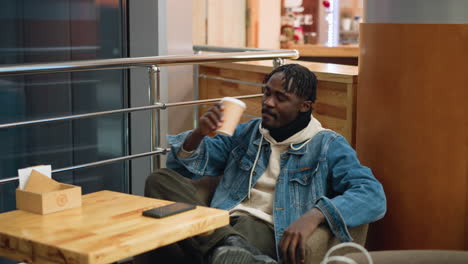 guy wearing denim jacket and beige hoodie sits comfortably with crossed legs in modern indoor caf , holding coffee cup while resting near table with smartphone and shopping bag, calm relaxed atmosphere