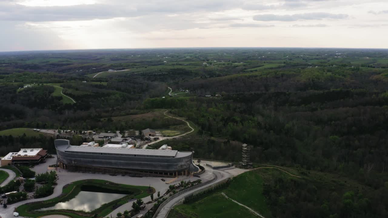 Drone shot of theme park in Kentucky shows massive ark-shaped structure surrounded by roads, buildings, and green hills. The unique attraction sits in wide natural landscape under cloudy sky.
