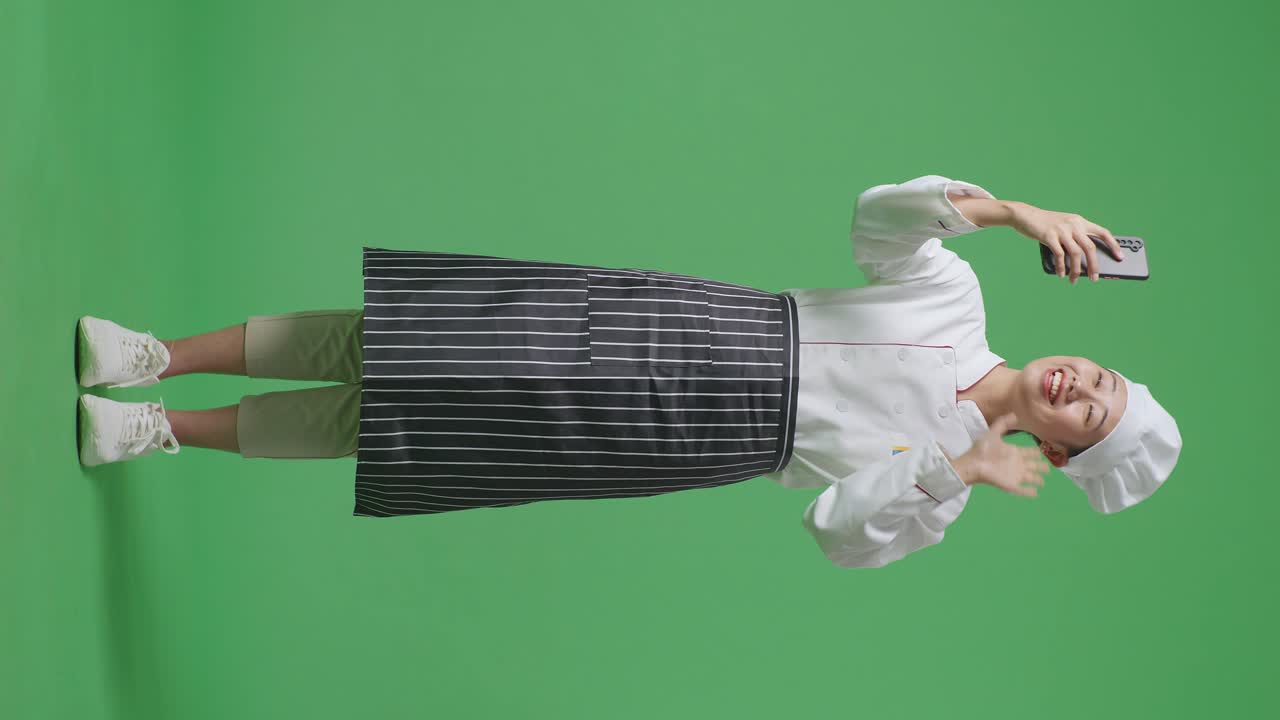 Full Body Of Asian Woman Chef Waving Hand Having A Video Call On Smartphone While Standing In The Green Screen Background Studio