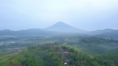 Revelar-Una-Toma-De-Un-Dron-Del-Paisaje-Rural-En-La-Mañana-Brumosa-Con-Una-Enorme-Montaña-En-El-Fondo