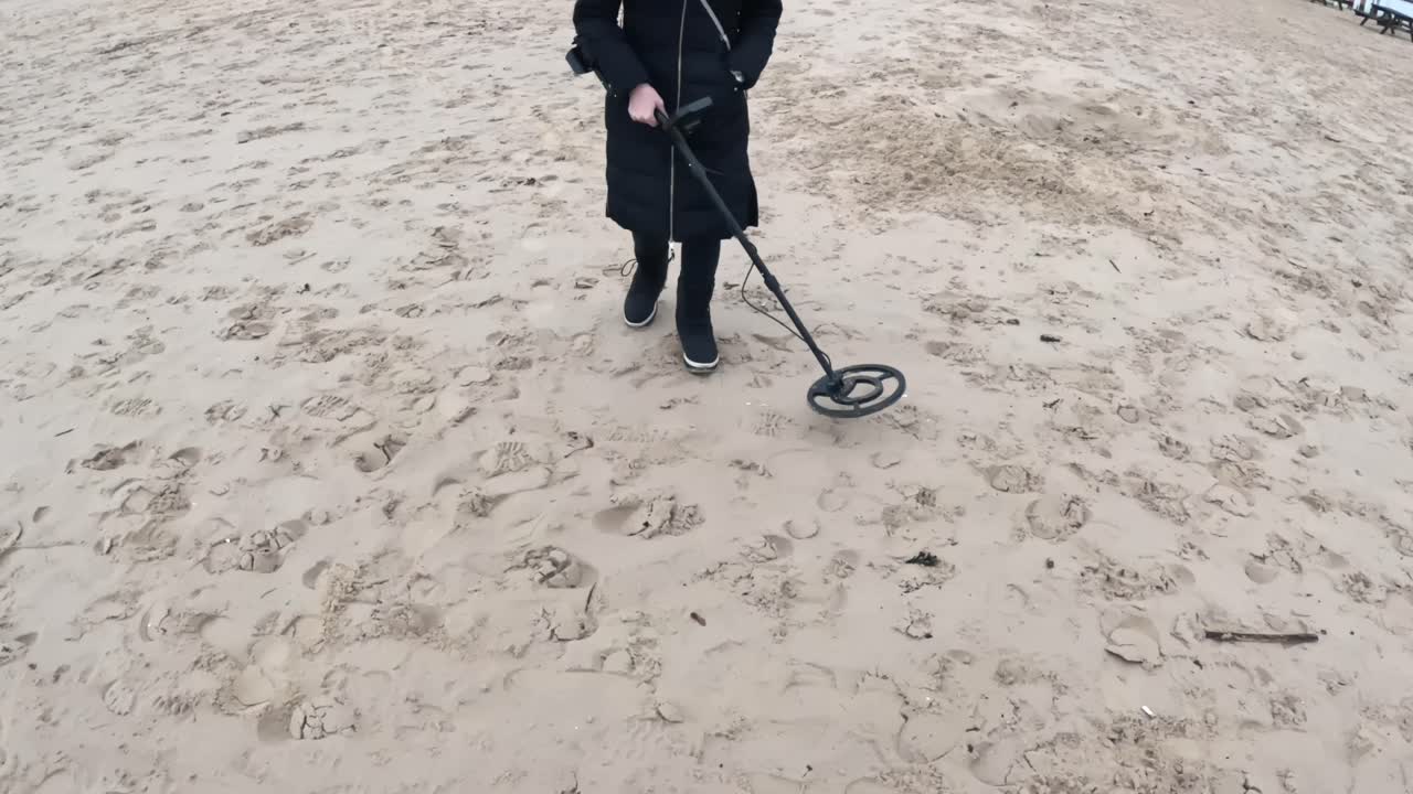 Persona che passa lentamente un metal detector sulla sabbia soffice della spiaggia, camminando tra le impronte alla ricerca di oggetti metallici nascosti