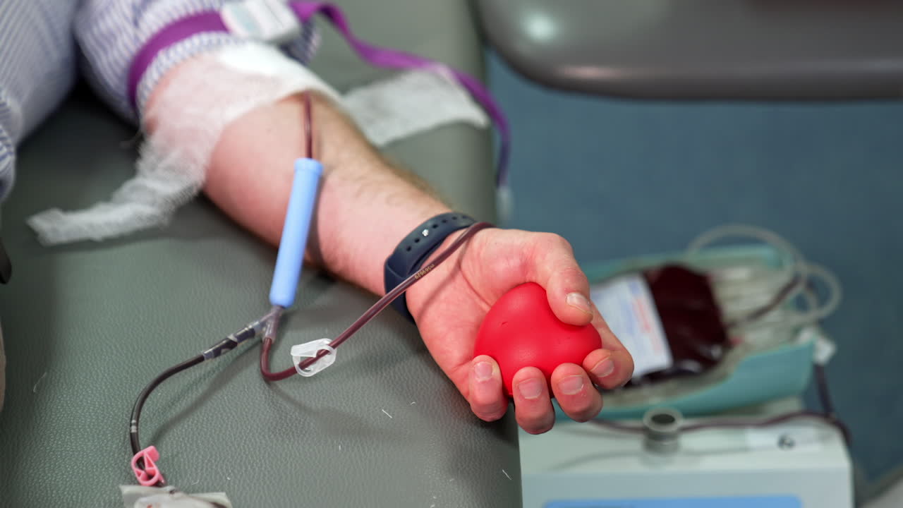 Red rubber heart is squeezed in male hand donating his blood. Close up. Plastic container with blood is on the apparatus at backdrop.