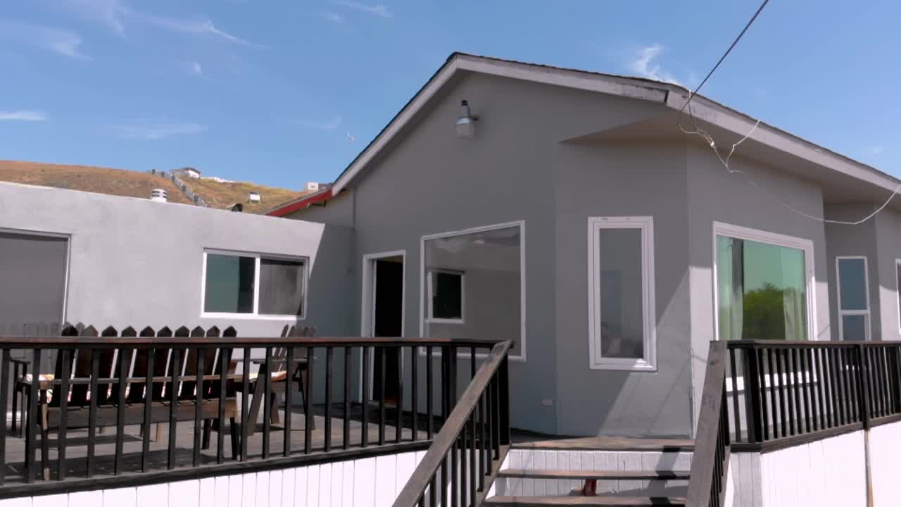 Exterior de casa moderna con barandilla de terraza y escaleras que conducen a la entrada, paredes limpias y grandes ventanales bajo cielo azul, mostrando diseño residencial y espacio de vida sencillo.