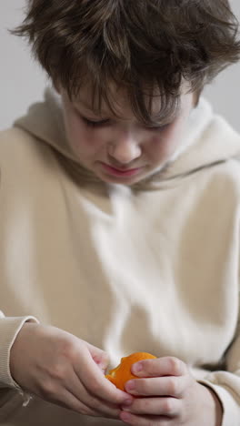 boy of twelve years old in beige hoodie peels a tangerine. kid wants to have a bite of a fruit. vertical video