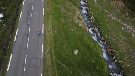 drone aerial following view of a single road cyclist climbing up an epic road whilst next to a small blue river stream surrounded by green grass