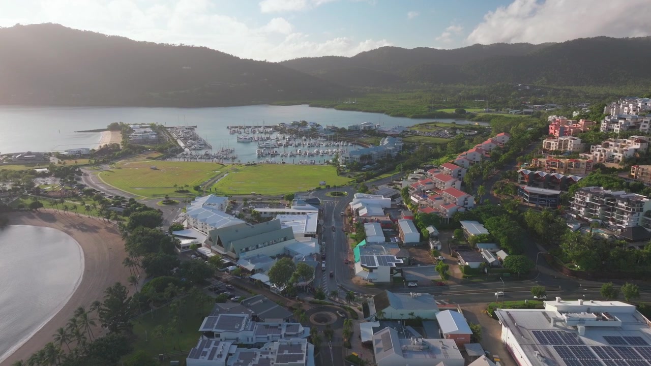 Cảnh quay drone trên không tại Đầm phá Vịnh Airlie Beach, trung tâm Rạn san hô Great Barrier, Whitsundays, Whitehaven, với nắng, mây, phố chính, ô tô, tòa nhà, sương mù, Biển San hô, bến du thuyền, cầu tàu, thuyền buồm, và chuyển động quay dần lộ ra