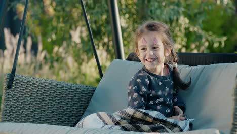 Portrait-of-a-cute-girl-with-face-painting-on-her-face,-resting-in-the-backyard-of-a-house-in-a-garden-swing