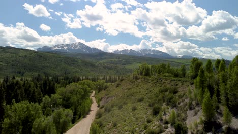 Camino-De-Tierra-Vacío-Que-Conduce-A-Las-Montañas-Con-Pinos-Y-Un-Río,-Aéreo