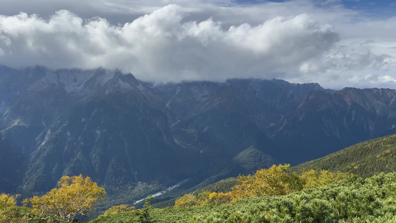 Pan shot across alpine shrubs toward the distant mountain ridges of the Northern Japanese Alps under a mix of bright sky and drifting clouds
