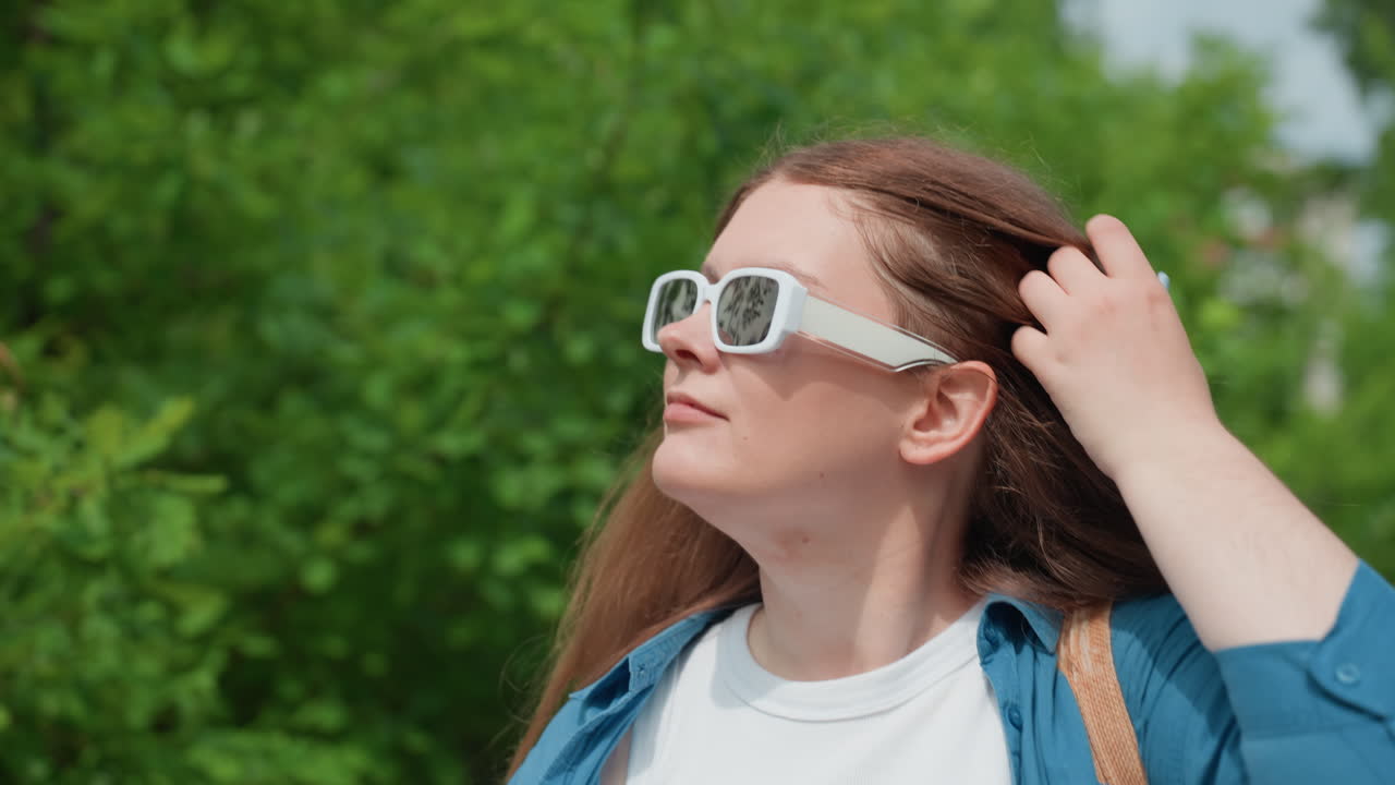 Крупный план: студент прогуливается в солнечный день в белых солнцезащитных очках и синей рубашке, поправляет волосы и наслаждается теплой погодой в окружении пышных зеленых деревьев и мягкого солнечного света