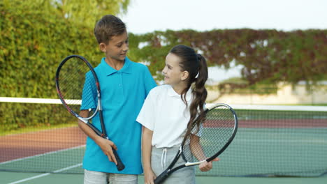 Retrato-De-Lindo-Hermano-Y-Hermana-Sosteniendo-Raquetas-Y-Sonriendo-Alegremente-A-La-Cámara-Mientras-Están-De-Pie-En-Una-Cancha-De-Tenis-Al-Aire-Libre-1