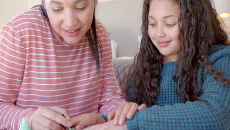 Happy-biracial-mother-sitting-and-painting-daughter's-nails-in-sunny-room