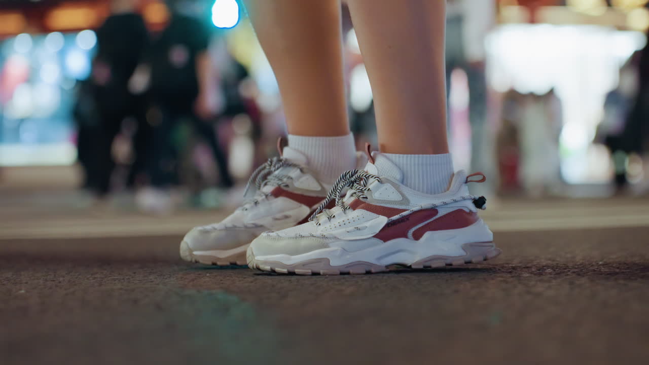 Primer plano de zapatillas en asfalto mojado bajo luces de neón, joven caminando entre una multitud difuminada, bokeh de calle urbana nocturna, pavimento reflectante, paseo casual rítmico, ambiente vespertino urbano.