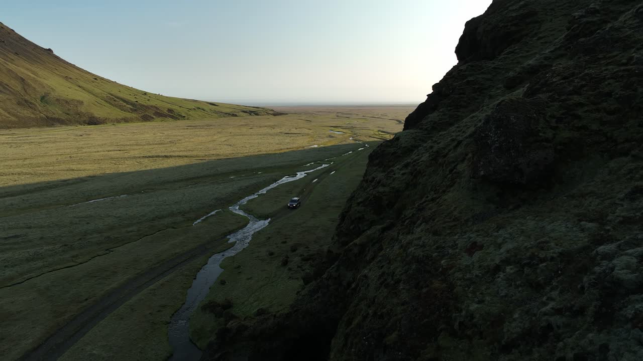 A lone off-road vehicle travels along a stream in a vast, remote valley in northern Iceland, bordered by steep cliffs and open plains.
