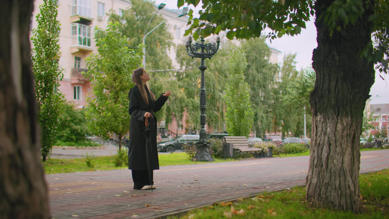 Woman in long black trench coat holding closed umbrella stands on tree-lined sidewalk in city park, looking upward with extended hand as if checking for raindrops, autumn leaves scattered on ground