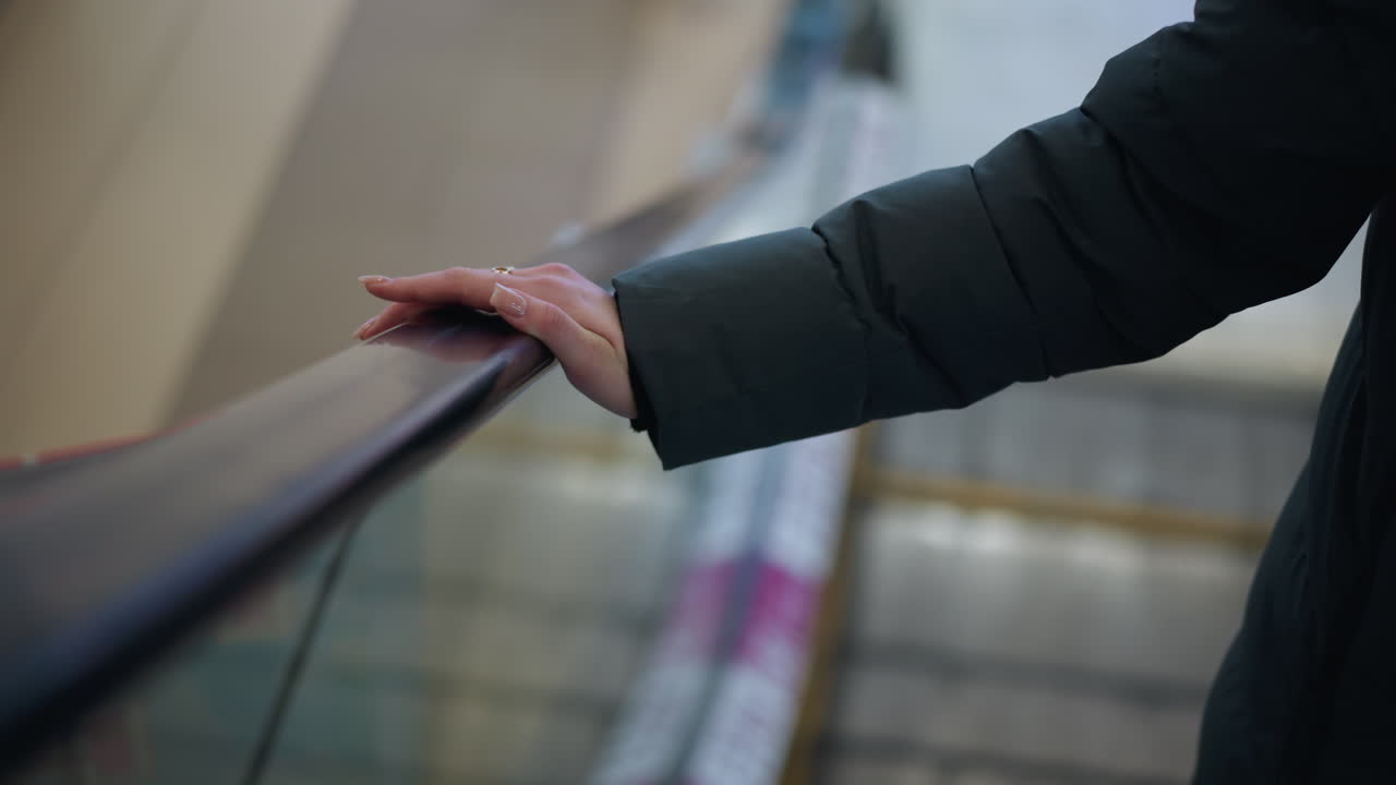primer plano de la mano con capucha negra descansando en el riel de una escalera mecánica en movimiento, anillos visibles en los dedos, el fondo presenta un reflejo con un borrón suave