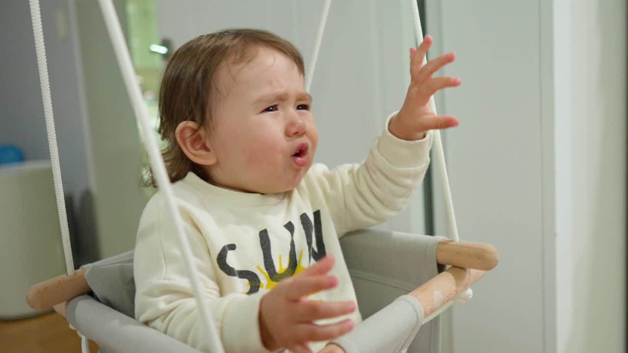 지루한 아기 소녀는 비명을 지르고 더 이상 출입구 그네에 앉아 울고 화난 얼굴을 보여주고 그녀를 데려가달라고 요청하고 싶지 않습니다.