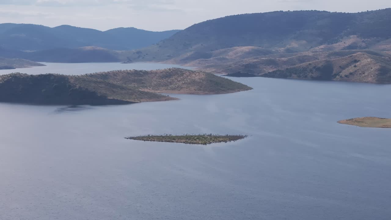 Drone orbit around small islands in the Cíjara reservoir, with golden hills and forested slopes in the background under a partly cloudy sky. Filmed in Badajoz, Spain, during late spring